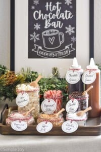 jars of toppings for Hot Chocolate Bar. Toppings are labeled on each jar and include mini marshmallows, peppermint sticks, chocolate chips, peppermint chips, toffee bits, and kosher salt. Bottles of chocolate sauce and caramel sauce sitting behind. Sign hanging behind says "Hot Chocolate Bar warm up buttercup"