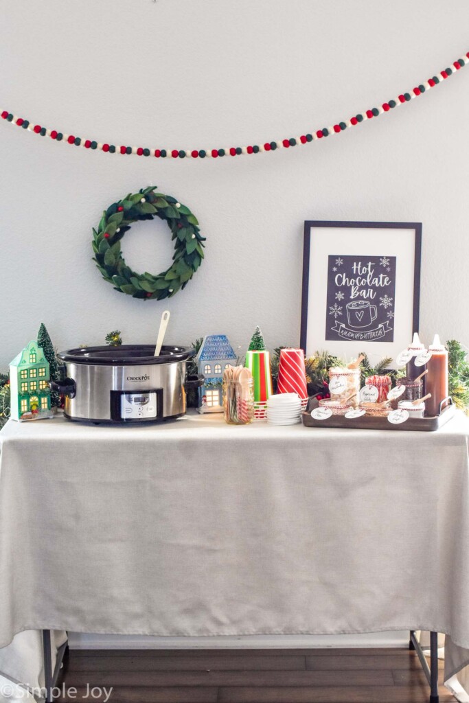 Table with crockpot of hot chocolate, cups, lids, wooden spoons, and toppings that make up a Hot Chocolate Bar