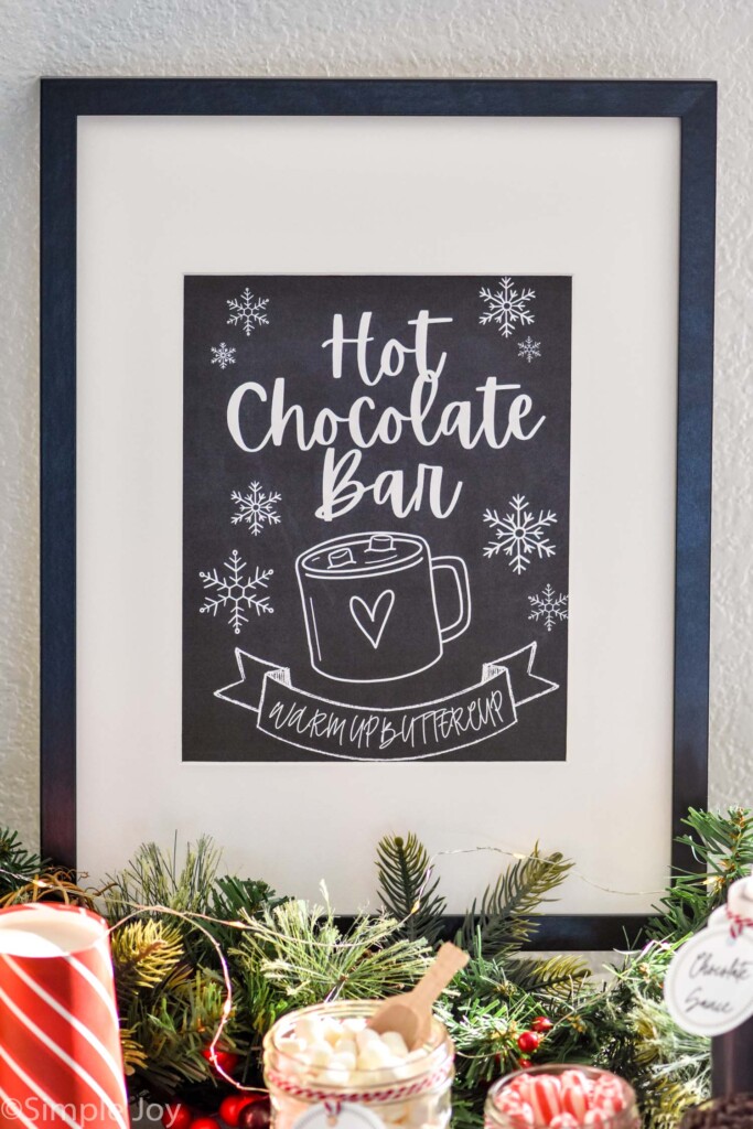 framed sign that says "hot chocolate bar warm up buttercup" with toppings for hot chocolate sitting in front.