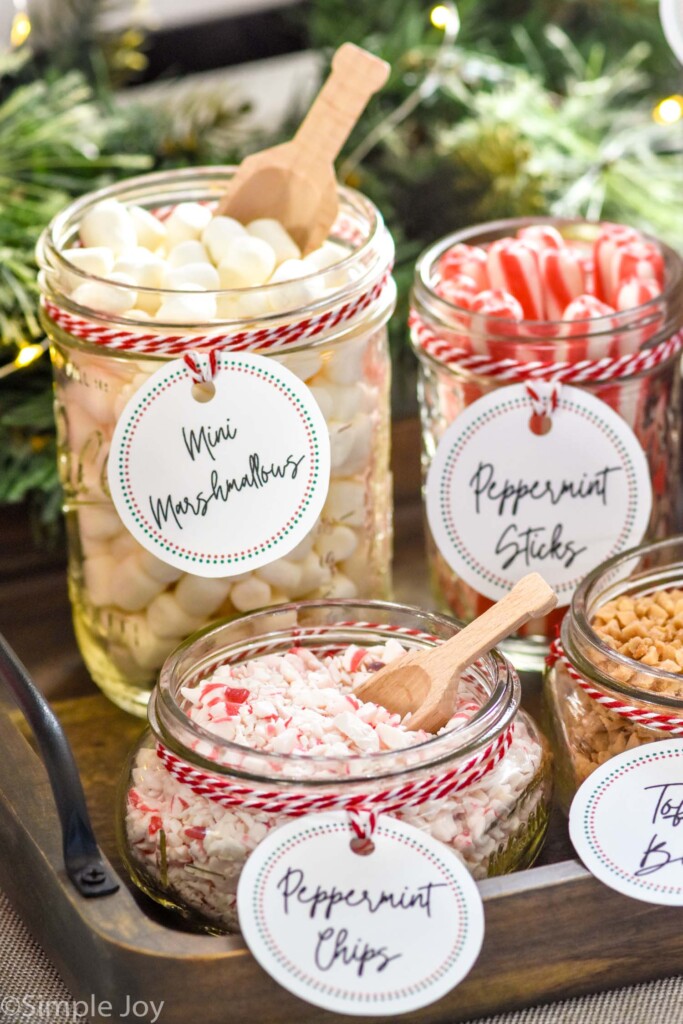 Jars of toppings and small wooden spoons for a Hot Chocolate Bar. Toppings include mini marshmallows, peppermint sticks, peppermint chips, and toffee bits, and are labeled on each jar.