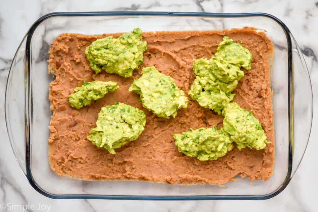 Overhead view of glass dish with beans and dollops of avocado mixture for seven layer dip recipe.