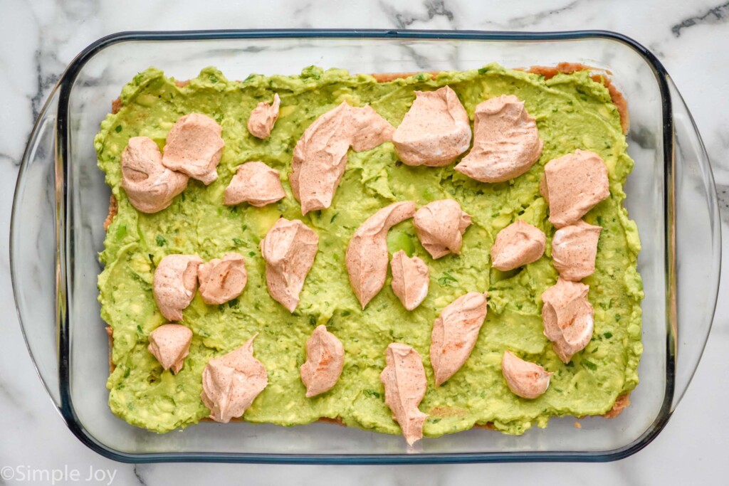 Overhead view of glass dish of avocado with dollops of seasoned sour cream for seven layer dip recipe.