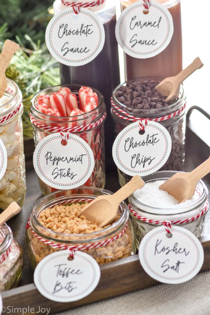 Jars of toppings for a Hot Chocolate Bar. Toppings are labeled on each jar and include mini marshmallows, peppermint sticks, chocolate chips, toffee bits, and kosher salt. Bottles of chocolate sauce and caramel sauce sitting behind.