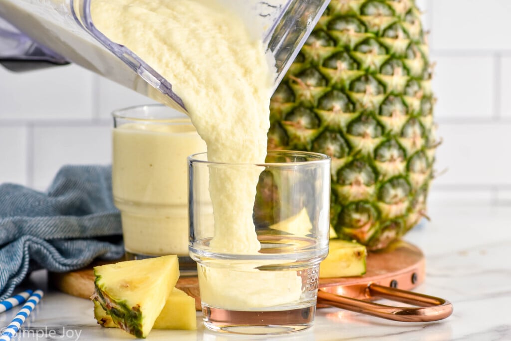 blender of Pineapple Smoothie pouring into a glass. Glass of Pineapple Smoothie, whole pineapple, and fresh pineapple wedges sitting in background