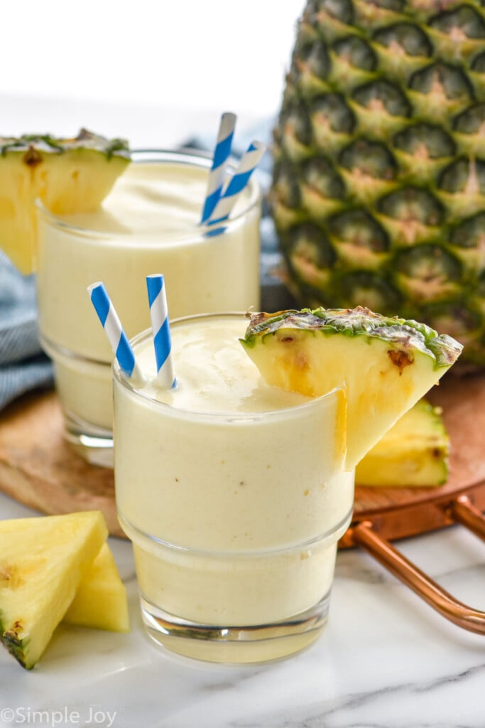 Two glasses of Pineapple Smoothie with two straws and a pineapple wedge on the rim. Fresh pineapple and pineapple wedges sitting in background