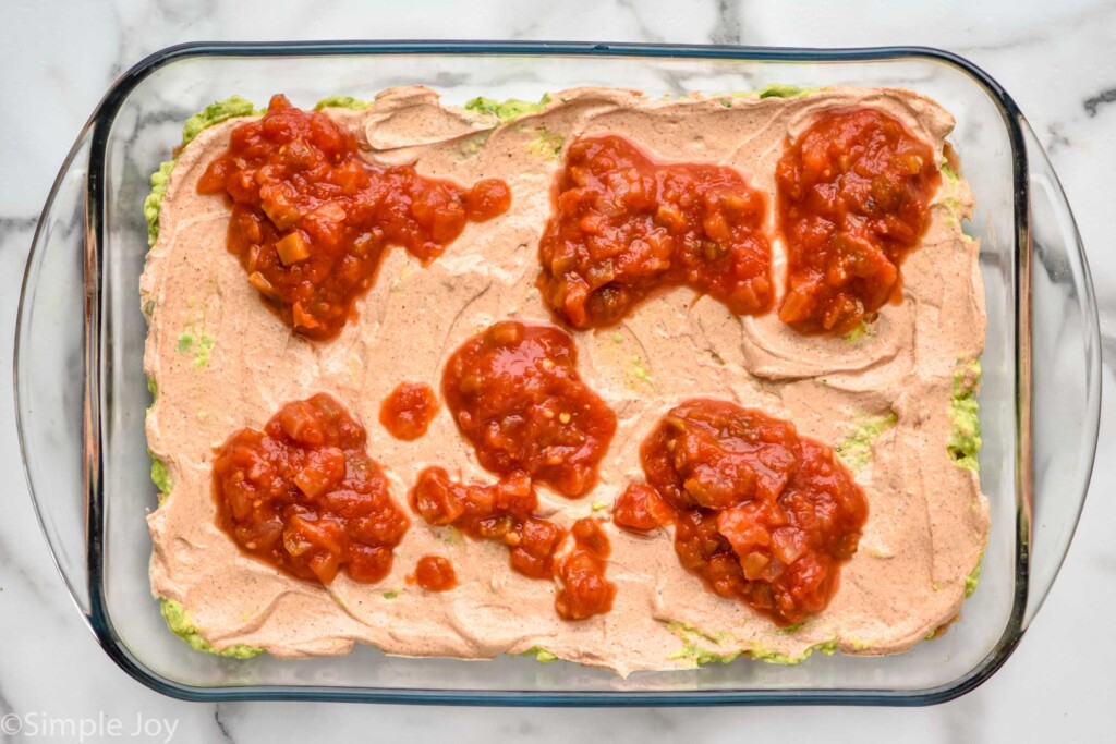 Overhead view of class baking dish topped with seasoned sour cream and dollops of salsa for seven layer dip recipe.