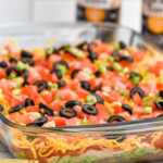 Side view of glass dish of Seven layer dip recipe topped with diced tomatoes, green onions, and black olives. Chips and bottles of beer beside.