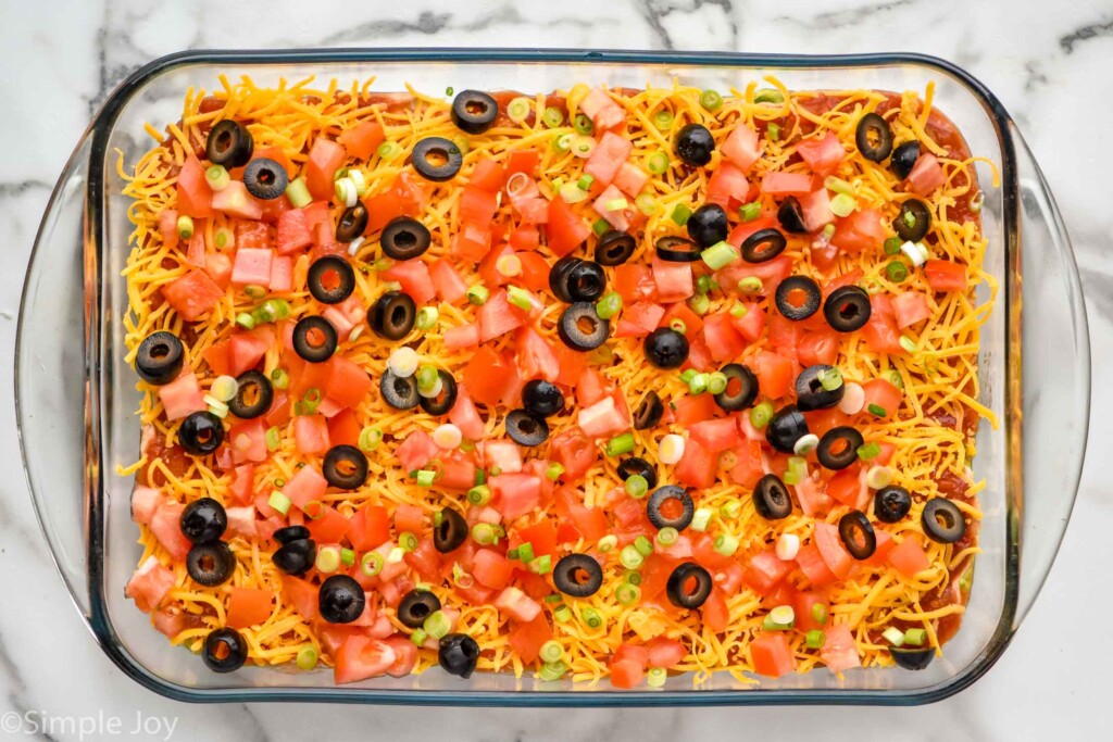 Overhead view of Seven layer dip in glass dish topped with diced tomatoes, green onions, and black olives.