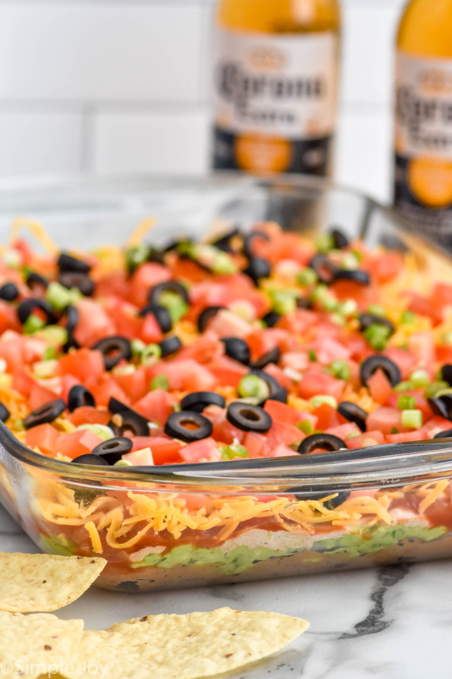 Side view of glass dish of Seven layer dip recipe topped with diced tomatoes, green onions, and black olives. Chips and bottles of beer beside.