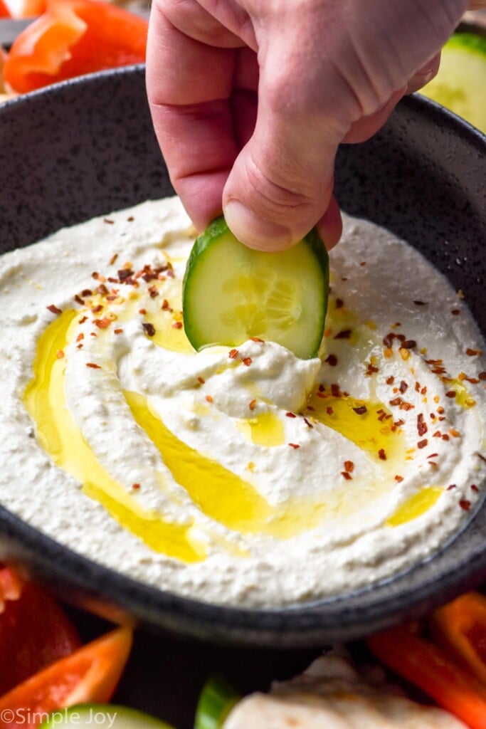 hand holding cucumber slice dipping into bowl of Whipped Feta Dip topped with olive oil and crushed red pepper flakes