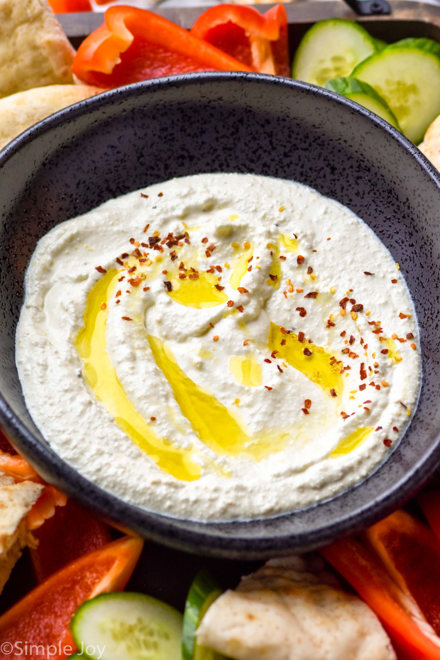 Bowl of Whipped Feta Dip topped with olive oil and red pepper flakes with cucumber slices, red pepper slices, and pita bread surrounding