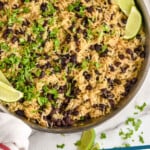 Pinterest graphic for Black Beans and Rice recipe. Image is overhead view of a skillet of Black Beans and Rice garnished with cilantro and lime wedges, tomatoes and wooden spoon beside. Text says, "Black Beans and Rice simplejoy.com."