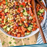 Pinterest graphic for chick pea salad recipe. Image is overhead view of a bowl of chick pea salad with spoons for serving, two drinks on counter beside. Text says, "chick pea salad simplejoy.com."