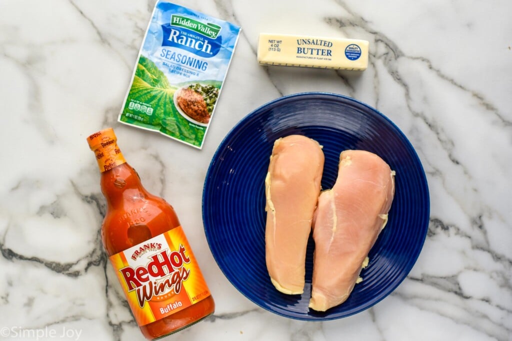 Overhead view of raw chicken on a plate with a bottle of buffalo sauce, a ranch seasoning packet, and butter beside for Slow Cooker Buffalo Chicken recipe.