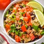 Overhead view of a bowl of Black Beans and Rice with diced tomatoes, cilantro, and lime wedges. Fork and tomatoes beside.