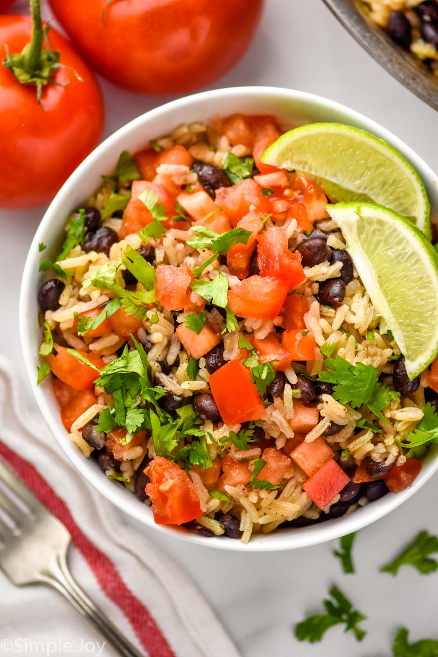 Overhead view of a bowl of Black Beans and Rice with diced tomatoes, cilantro, and lime wedges. Fork and tomatoes beside.