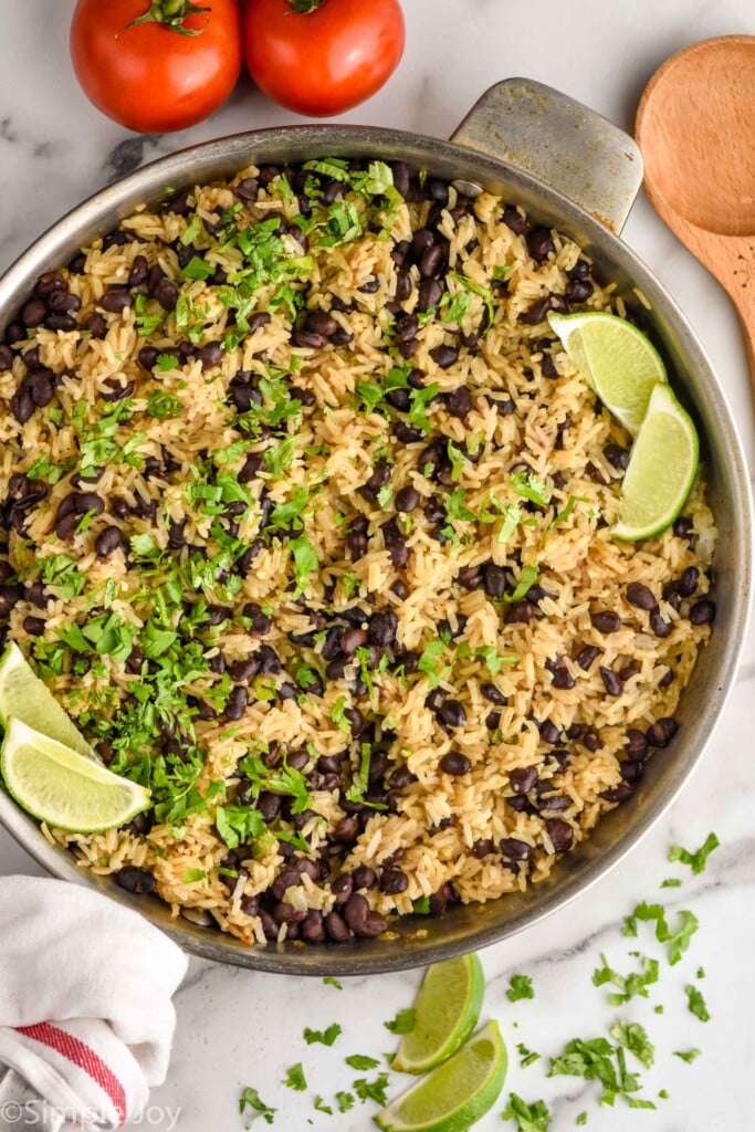 Overhead view of a skillet of Black Beans and Rice garnished with cilantro and lime wedges, tomatoes and a wooden spoon beside.