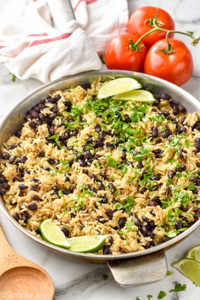 skillet of Black Beans and Rice recipe garnished with lime wedges and cilantro. Tomatoes and a wooden spoon on counter beside