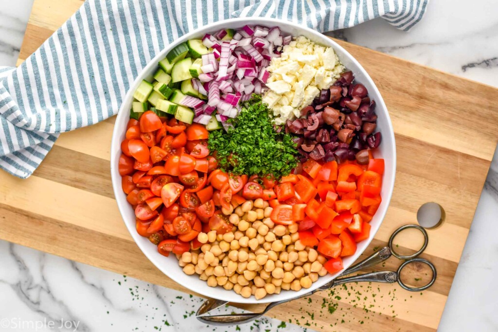 Overhead view of a large bowl with chopped ingredients for chick pea salad recipe. Tongs beside bowl for mixing.