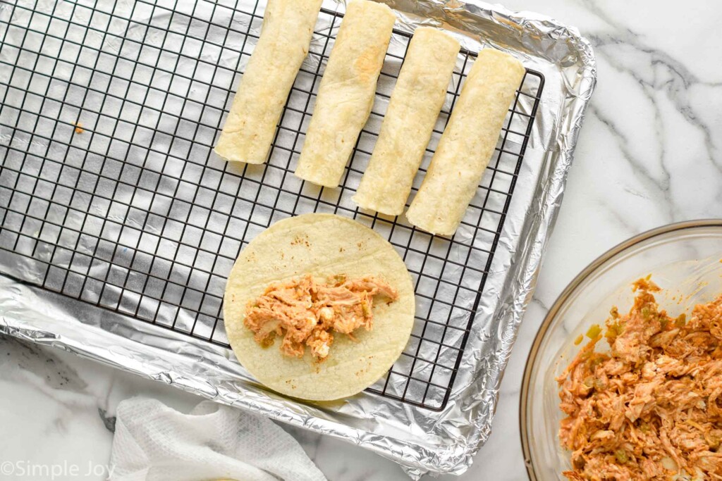 Overhead view of baking sheet with wire rack of Chicken Taquitos in progress. Bowl of Chicken Taquitos filler beside for recipe.