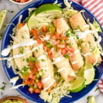 Overhead view of a plate of Chicken Taquitos on a bed of shredded lettuce, garnished with pico de gallo, lime wedges, and cilantro. Lime wedges, pico de Gallo, and guacamole beside plate.