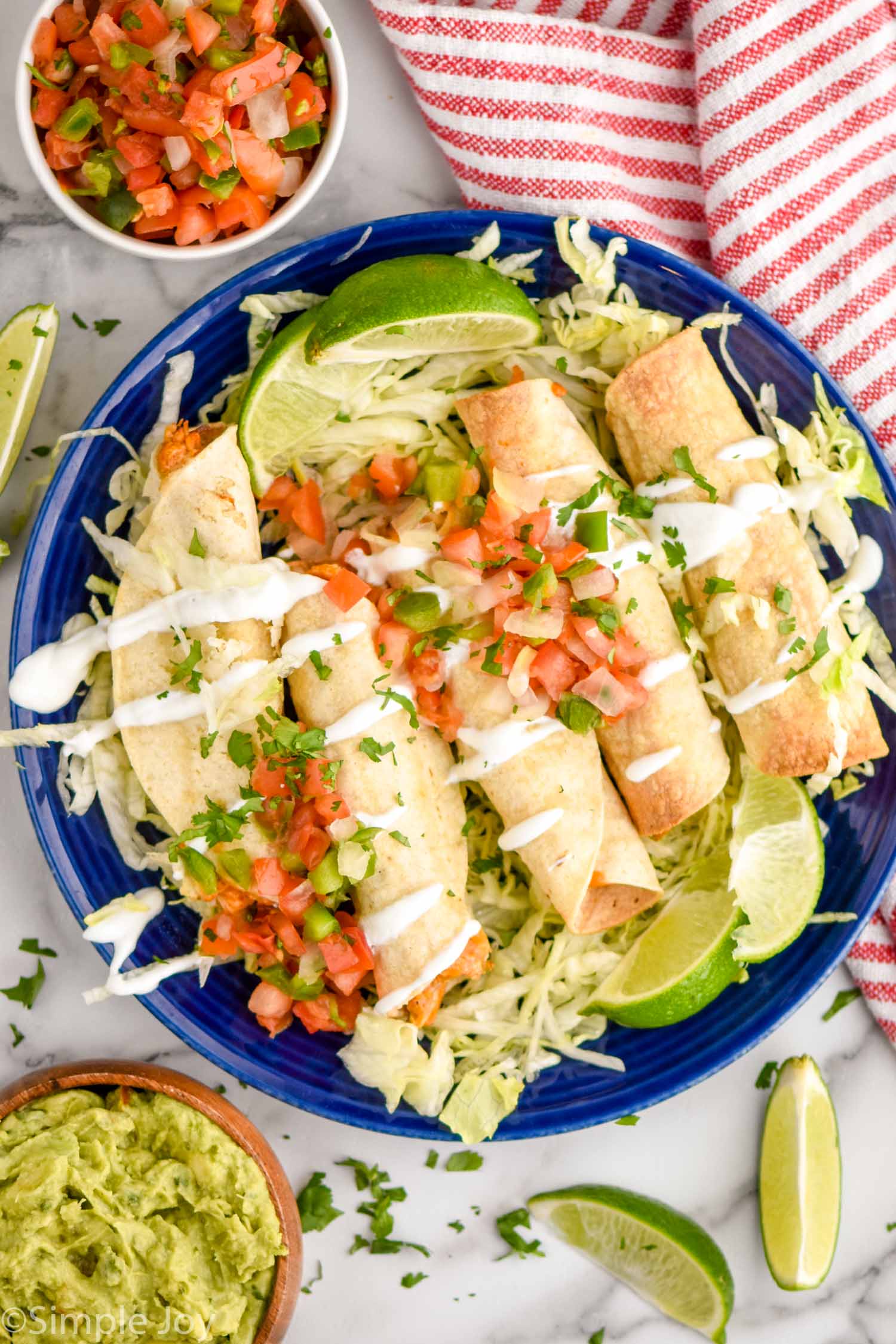 Overhead view of a plate of Chicken Taquitos on a bed of shredded lettuce, garnished with pico de gallo, lime wedges, and cilantro. Lime wedges, pico de Gallo, and guacamole beside plate.