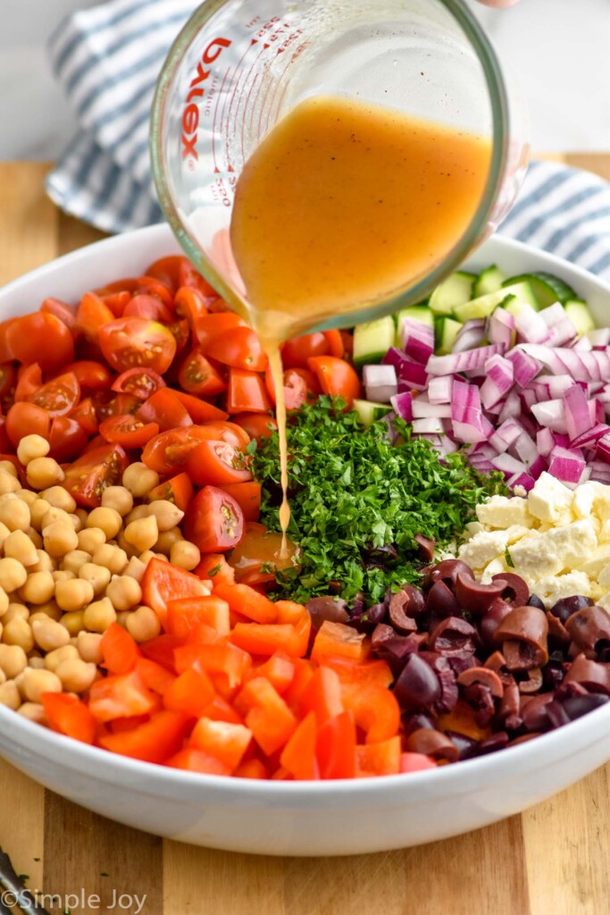 Side view of glass measuring cup of dressing being poured onto a bowl of chopped ingredients for chickpea salad recipe.