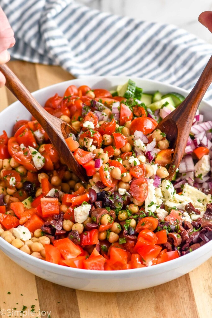 Person's hands holding spoons in bowl of chickpea salad for serving. Salad is garnished with parsley