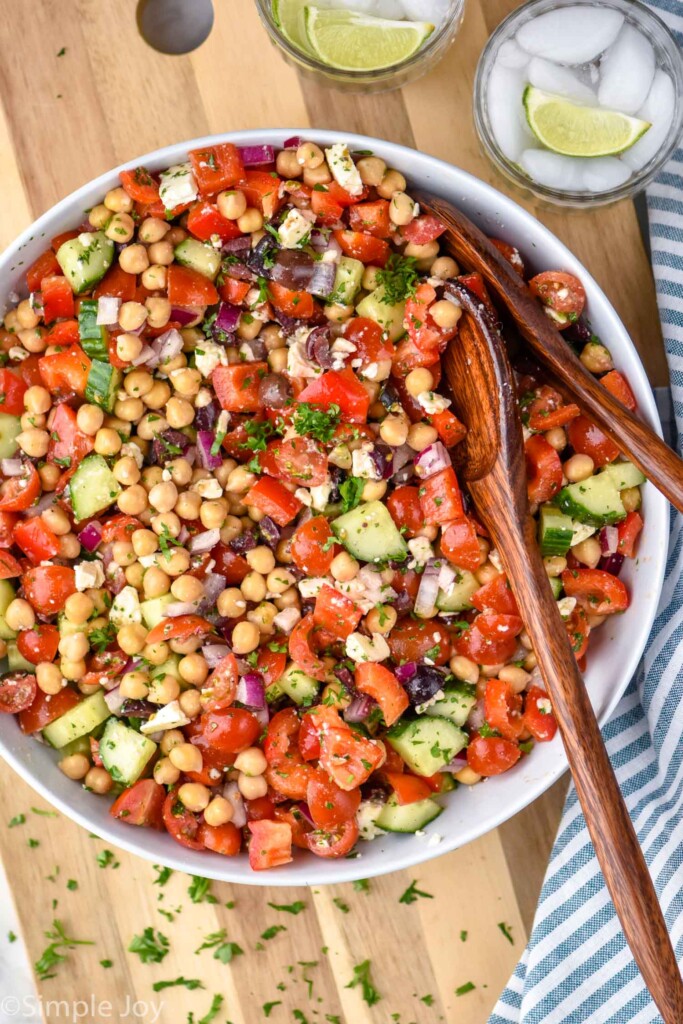 Overhead view of a bowl of chickpea salad garnished with parsley with spoons for serving. Beverages on counter beside.