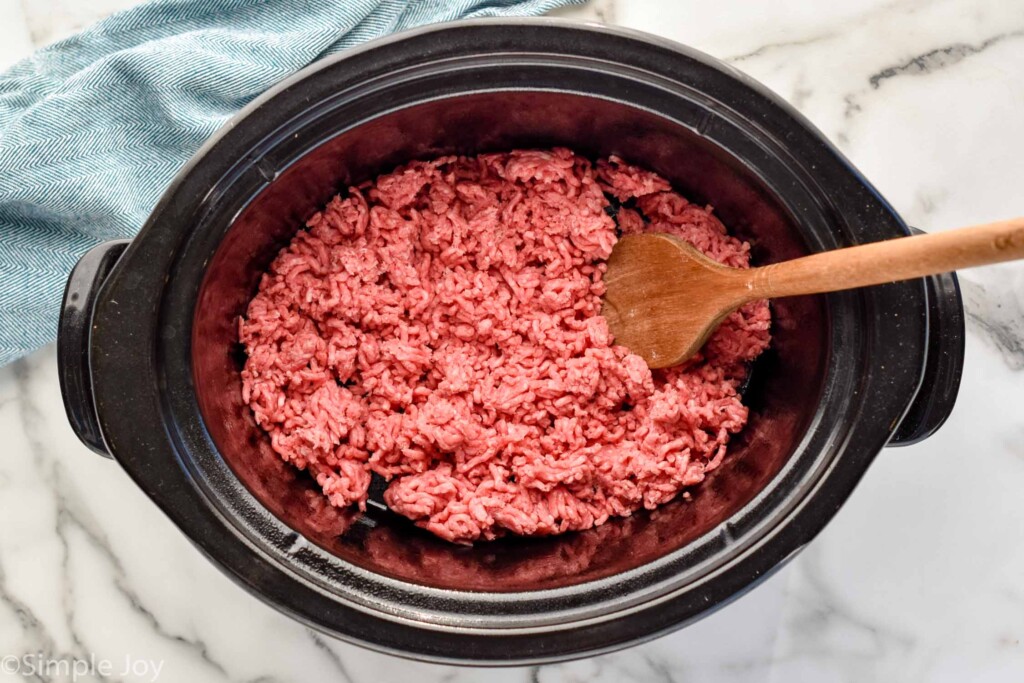 Overhead view a crock pot with hamburger and a wooden spoon for Slow Cooker Taco Meat recipe