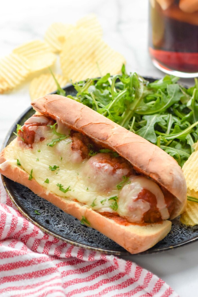 meatball sub served on a plate with salad and potato chips. Potato chips and iced tea beside.