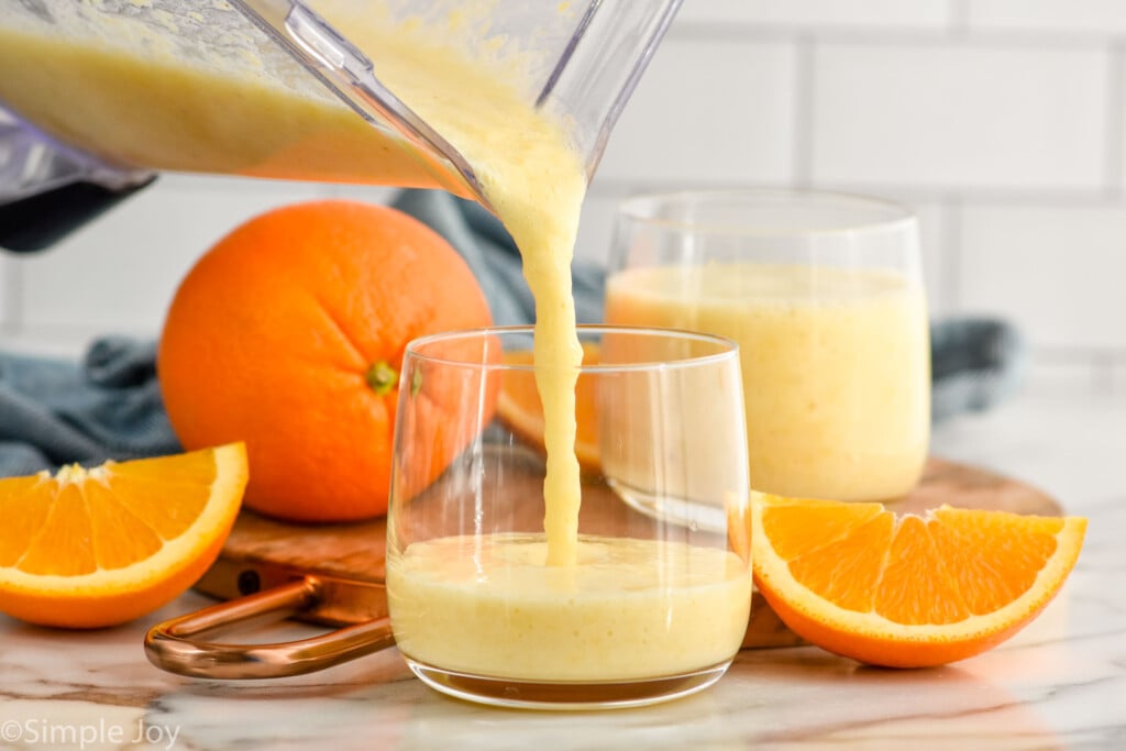 Side view of a blender pitcher of orange smoothie being poured into glasses. Orange and orange slices on counter beside for garnish.