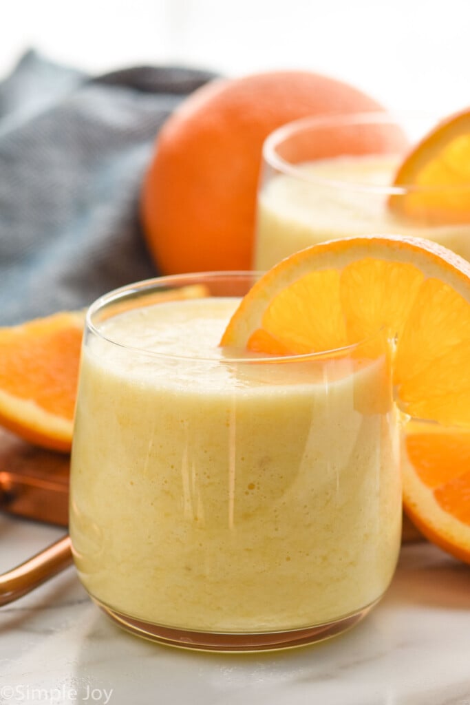 Side view of glasses of orange smoothie with orange slices on the rim. Orange and orange slices beside for garnish.