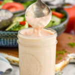 Side view of a jar of Thousand Island Dressing with a spoon scooping some out of the top. A salad in a bowl is in the background.