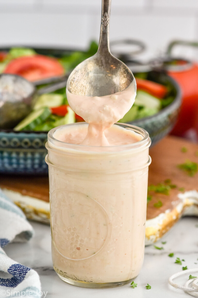 Side view of a jar of Thousand Island Dressing with a spoon scooping some out of the top. A salad in a bowl is in the background.