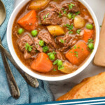 Pinterest graphic for Crockpot Beef Stew recipe. Overhead view of a bowl of Crockpot Beef Stew with silverware, bread, and a glass of red wine beside. Text says, "crockpot beef stew simplejoy.com."