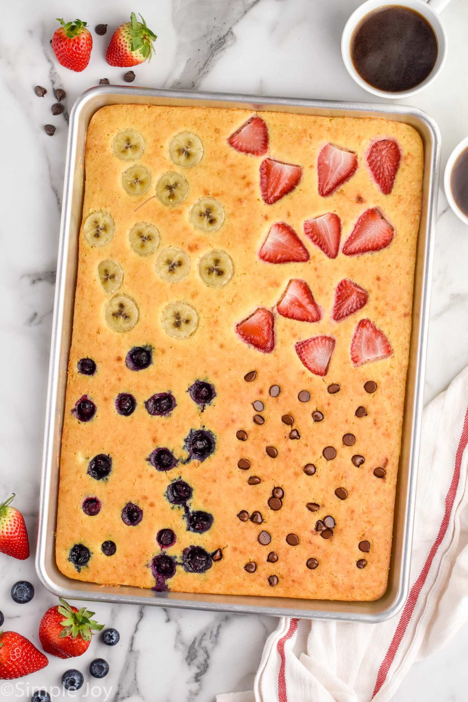 Overhead view of Sheet Pan Pancakes garnished with bananas, strawberries, blueberries, and chocolate chips in quadrants. Cup of coffee and berries beside.