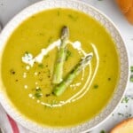 Overhead view of a bowl of Asparagus Soup garnished with cream and two pieces of asparagus, bread beside