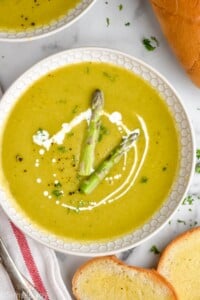 Overhead view of a bowl of Asparagus Soup garnished with cream and two pieces of asparagus, bread beside