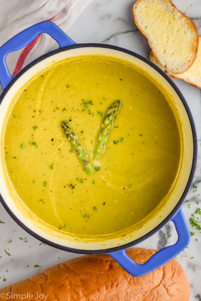 Overhead view of a pot of Asparagus Soup garnished with two pieces of asparagus, bread beside.