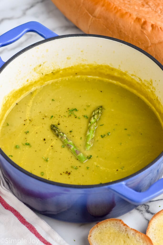 Pot of Asparagus Soup garnished with two pieces of asparagus, bread beside