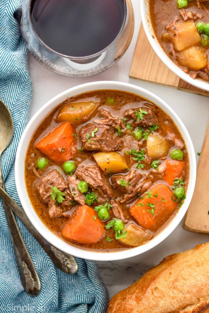 Overhead view of a bowl of Crockpot Beef Stew with silverware, bread, and a glass of red wine beside.