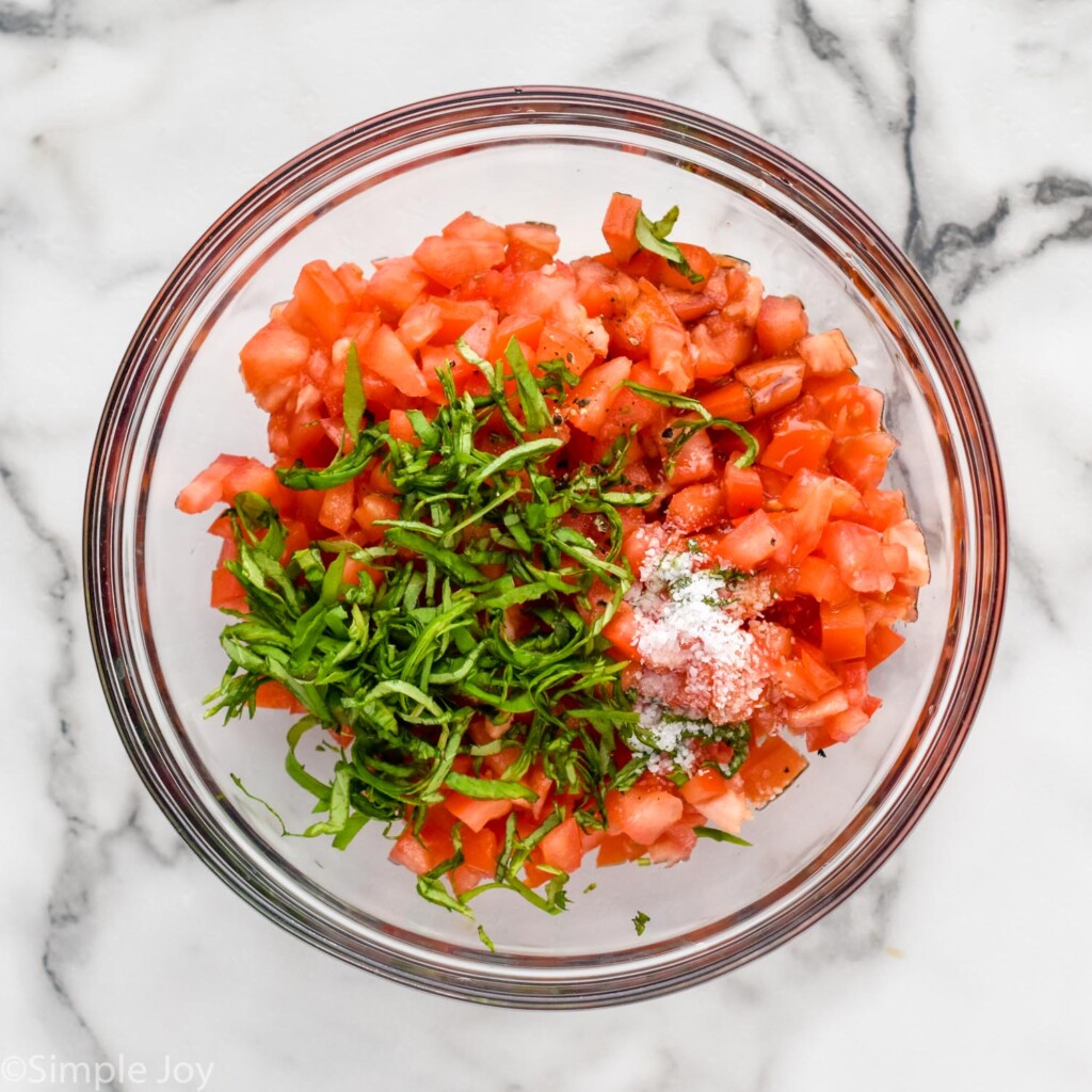 Overhead view of a mixing bowl of diced tomatoes, salt, and sliced basil leaves for Bruschetta recipe.