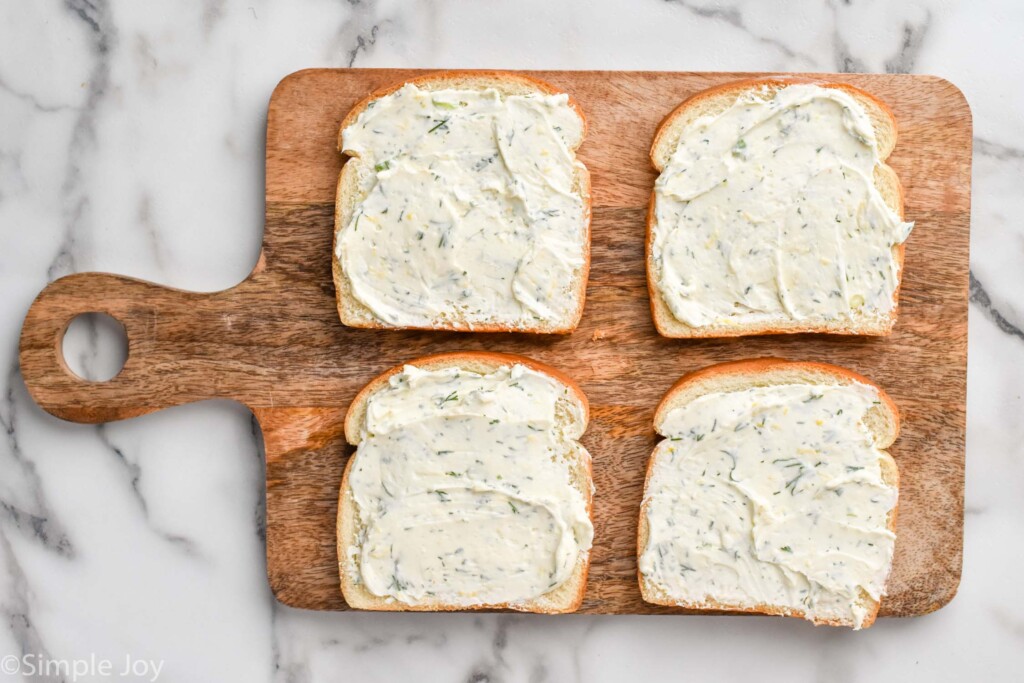 Overhead of wooden cutting board with four slices of bread topped with cream cheese filling mixture to make cucumber sandwiches