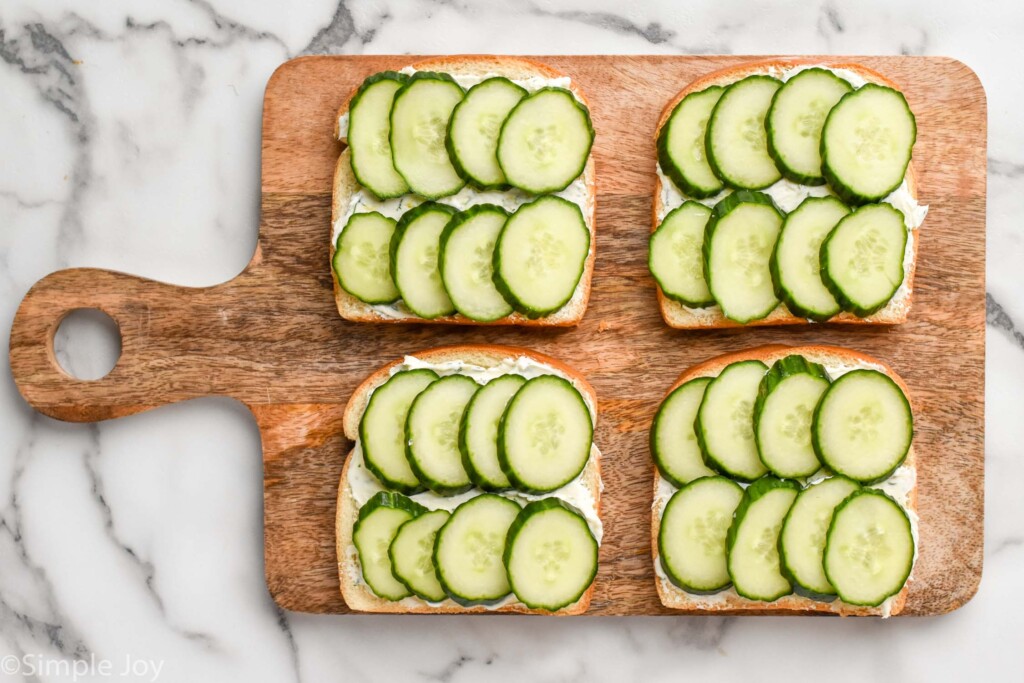 Overhead of wooden cutting board with four slices of bread topping with cucumber sandwich ingredients to make cucumber sandwiches