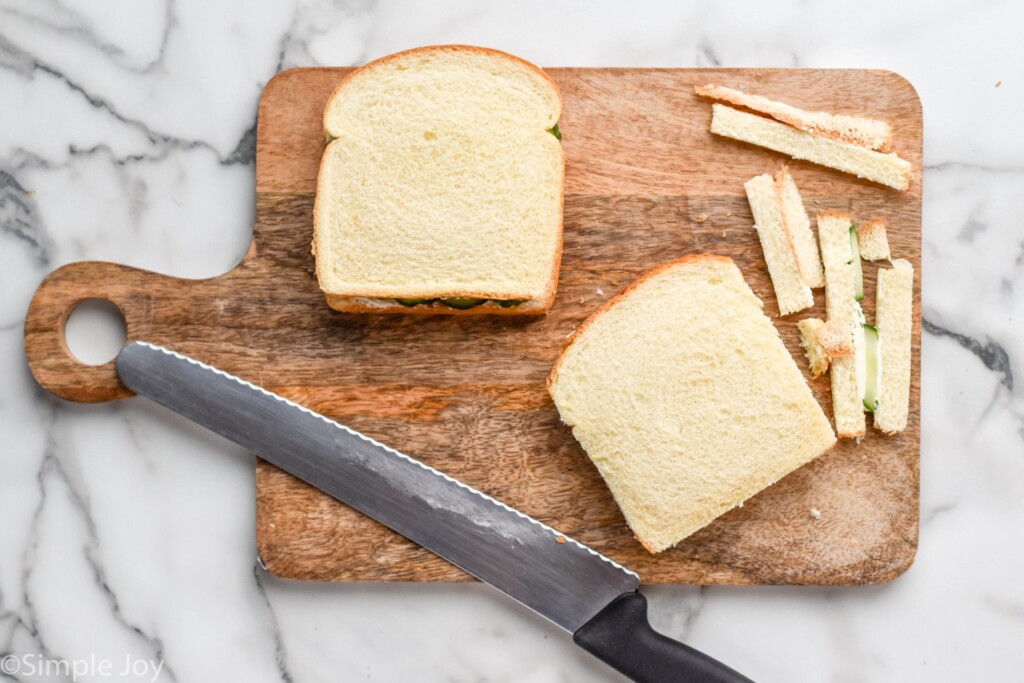 Overhead of wooden cutting board with two cucumber sandwiches, one with crust cut off and pieces sitting beside, large serrated knife also sitting beside.