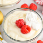 Bowl of Pudding garnished with raspberries and whipped cream, raspberries and spoons beside.