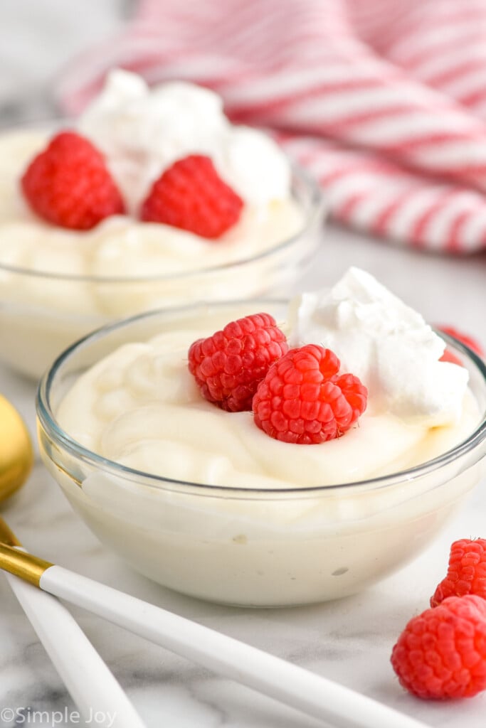 Bowls of Pudding garnished with raspberries and whipped cream, raspberries and spoons beside.