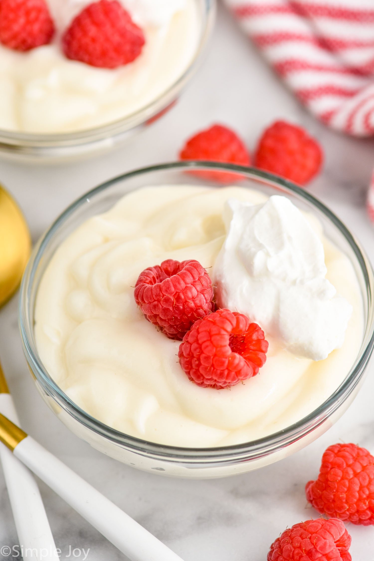 Bowl of Pudding garnished with raspberries and whipped cream, raspberries and spoons beside.