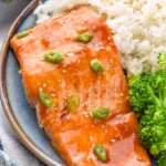 Teriyaki Salmon garnished with green onions and sesame seeds, served on a plate with white rice and broccoli. Fork beside.